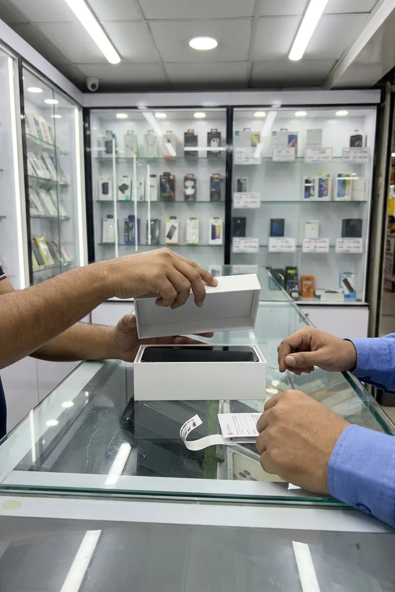 A shopkeeper lifts the lid off a new phone’s box on a glass counter, its receipt beside it, as the customer looks on
