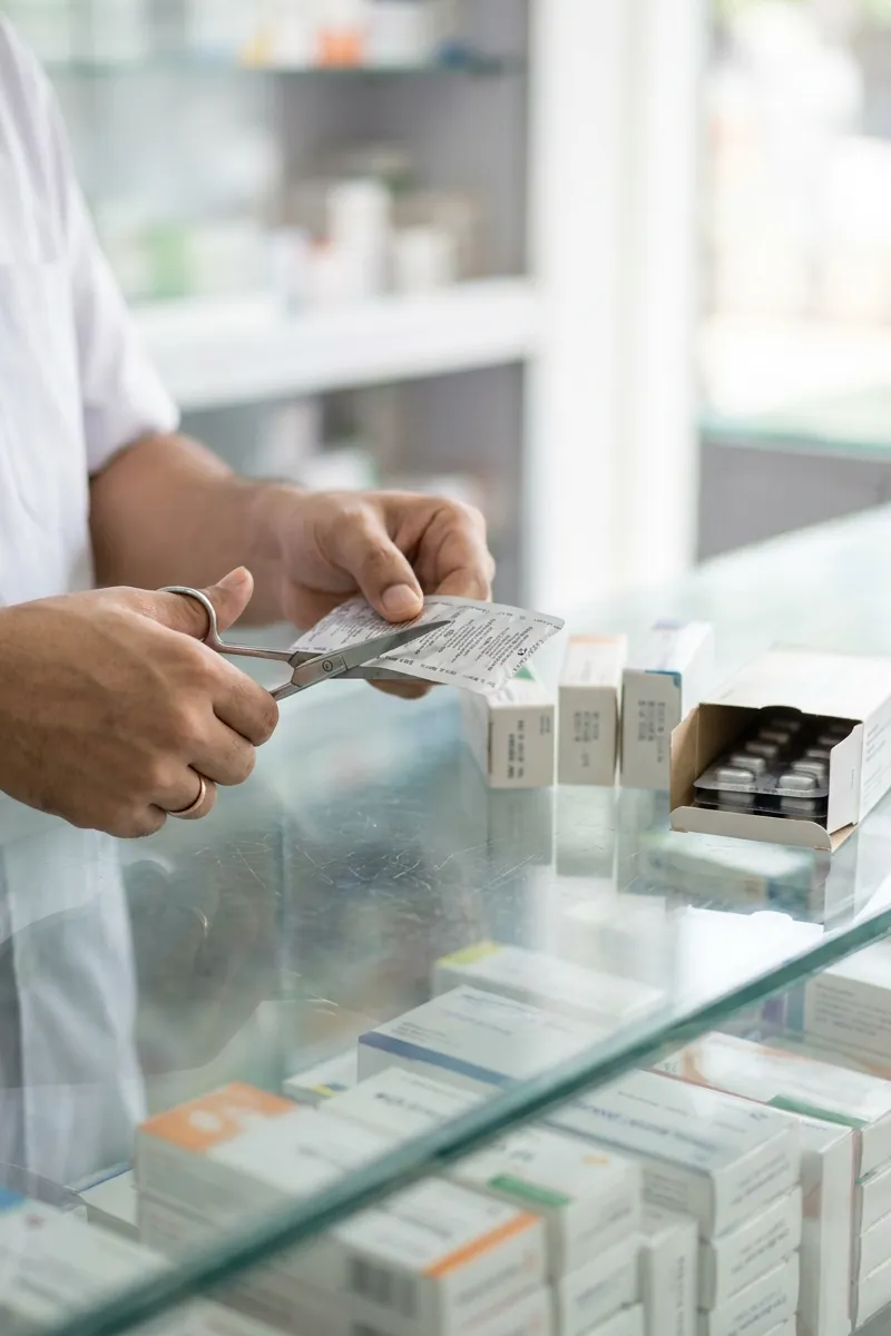 A pharmacist snips tablets off a blister strip with scissors at a glass counter, an open box of strips beside him